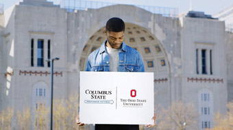 Columbus State student holding preferred pathway sign (Columbus State to Ohio State) in front of the Ohio State University's Horseshoe stadium.