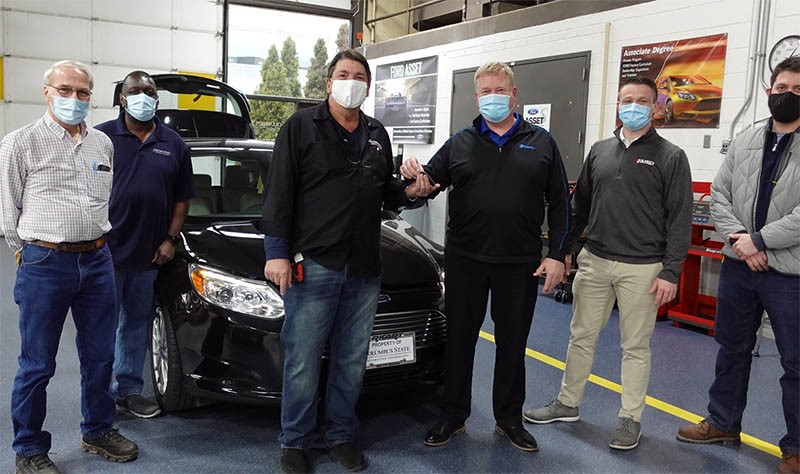 A picture of the vehicle being donated with professors and some Ricart employees standing in front of it. 