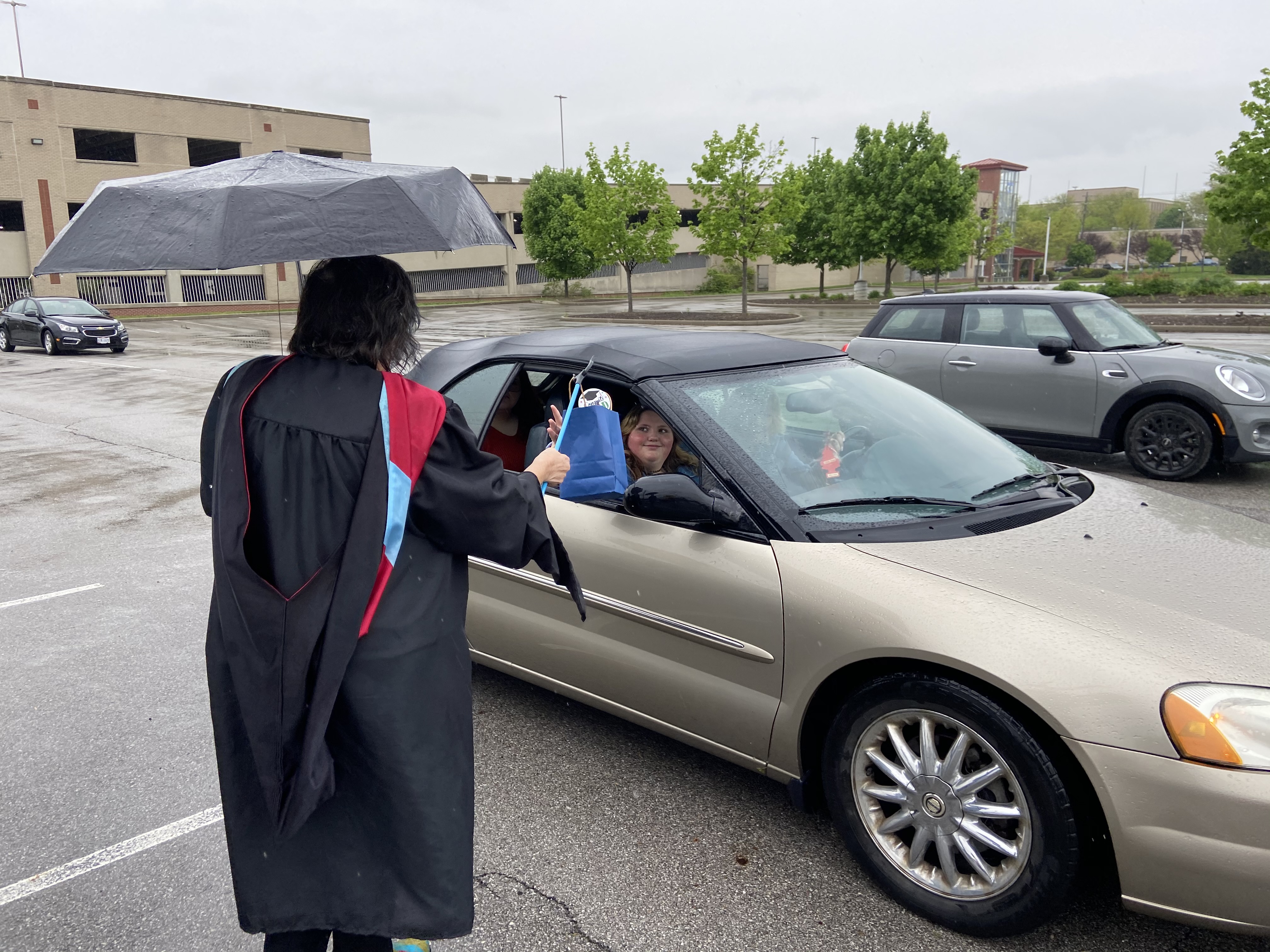 A picture of a staff or faculty member handing a diploma to a graduate in their car. 